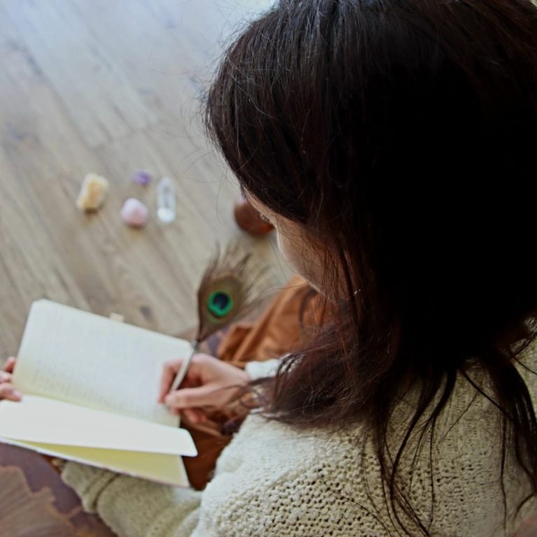 mentorías Conciencia de Corazón Persona escribiendo en un cuaderno, rodeada de piedras y plumas sobre el suelo.