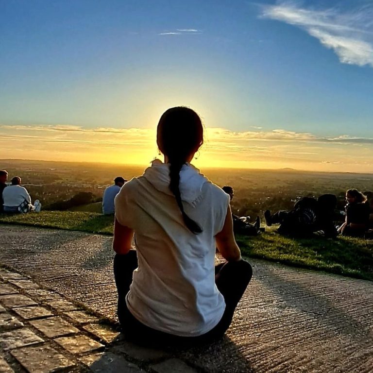 Meditaciones Conciencia de Corazón Persona sentada al atardecer, observando un paisaje con otras personas al fondo.