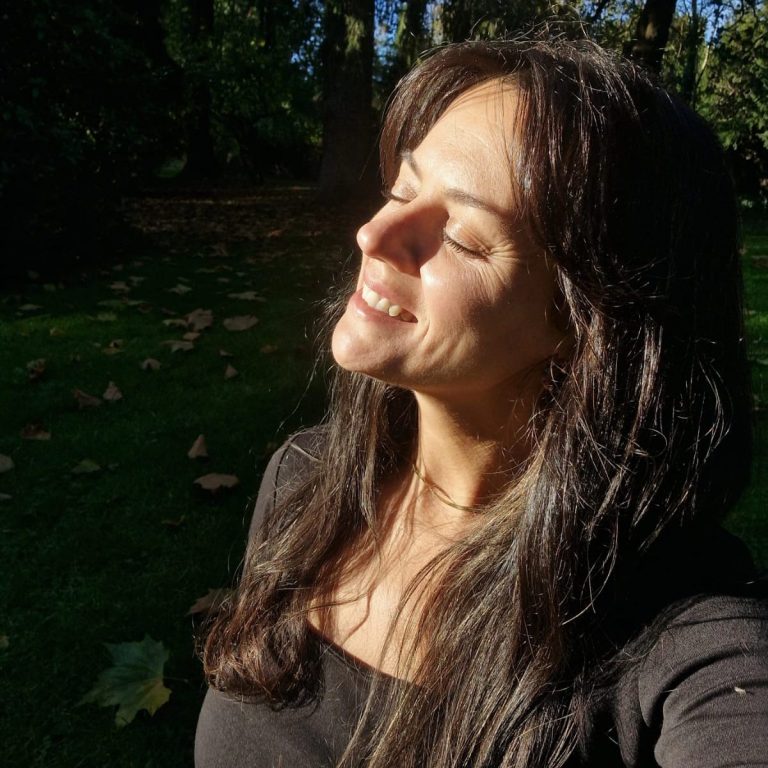 Fundadora de Conciencia de Corazón Mujer sonriendo al sol en un entorno natural con luz suave y hojas alrededor.